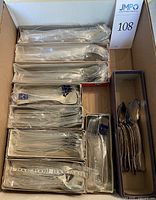 Box containing multiple individually wrapped stainless steel spoons packaged in cardboard trays, also includes loose silverplated teaspoons