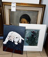 Three paintings grouped: portrait in larger frame, floral in white frame, puppy on canvas leaning in front.
