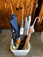 Photo showing 8 umbrellas standing inside a white plastic bin. Four tall umbrellas have wooden curved handles and vary in fabric colors. Four compact foldable umbrellas visible in the bin.
