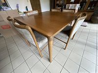 Diagonal view of oak dining table with six matching chairs surrounding it, showing the wood finish and chair upholstery.