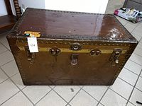 Front view of a large rectangular brown vintage luggage trunk with metal latches, lock, and riveted edges. Shows some wear and tear.