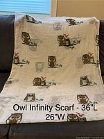 Cream infinity scarf with owl pattern laid out on couch, showing full width and length dimensions written on the scarf.