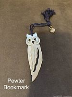 Photo of pewter owl-shaped bookmark showing detailed wings, cut-out eyes, and navy blue braided tassel with a small tag.