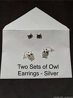 Two sets of owl silver earrings displayed on a white card, including one pair of owl studs and one pair of owl dangle earrings.