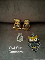 Four owl sun-catchers arranged on a dark fabric surface, two gold-tone owls with clear faceted crystal bodies perched on bars, one silver filigree owl with crystal body, and one colorful enamel owl with number '24' tag.