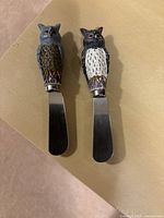Photo showing the two owl-themed serving spreaders with detailed handles and stainless blades on a beige background.