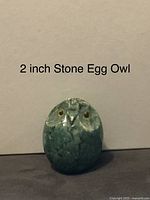 Two inch stone egg carved and painted as an owl head, showing mottled green texture and detailed owl face.