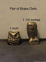 Two small solid brass owl figurines shown side by side with measurement labels. One owl is stout and square-shaped about 1 inch wide, the other owl is taller with detailed surface texture about 1.5 inches tall.