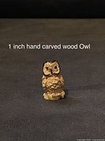 Close-up photo of a 1 inch hand carved wooden owl figurine sitting on a dark surface, showing detailed feather carving and natural wood color.