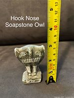 Photo showing one soapstone owl sculpture next to measuring tape indicating approximately 4 inches tall. The owl has a unique hook nose carved face with engraved eyes and mouth details.