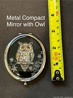 Top view of one oval metal compact mirror with painted owl design and measuring tape showing size about 2.5 inches