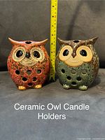 Photo of two ceramic owl-shaped candle holders, one red and one green, positioned side by side with a measuring tape behind indicating height approximately 5.5 inches.