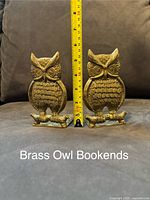 Photo of two brass owl bookends placed side-by-side on a couch with a measuring tape showing height of approximately 5.5 inches