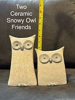 Photo showing two ceramic snowy owl figurines with a measuring tape indicating approximately 8 inches tall, placed on a surface with a dark background.