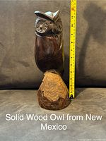 Front view of a solid carved wood owl sculpture next to a measuring tape showing about 10 inches tall. Brown wood with carved details and a natural finish.
