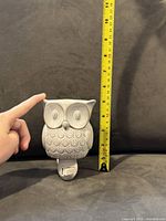 Close-up photo of ceramic owl air freshener against a black background measured with a yellow tape showing about 6 inches height, fingers for scale.