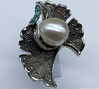 Front view showing the textured leaf petal design with the central white baroque pearl and a line of four emerald stones on one petal, surrounded by marcasite stones.