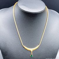 Front view of the gold-plated necklace on a black bust, showing the chain, pendant with textured gold segments, small white gem in a heart shape, and green teardrop stone.