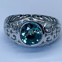 Front view of sterling silver ring with round faceted bluish-green sapphire in bezel setting, Art Nouveau-style filigree band.