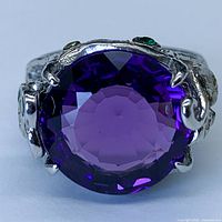 Front close-up of large round violet amethyst gemstone set in sterling silver band with white gold plating and small green and blue accent stones.