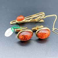 Front angled view of amber cufflinks and matching tie clip showing the oval amber stones set in gold-tone metal on a black background.