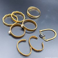 Photo showing seven gold-tone bracelets on a black background, different styles including solid bands, bangles, pearl accented cuffs, and flexible bracelet with black adornments.