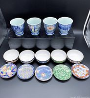 Five Arita porcelain tumblers in various traditional Japanese patterned designs, atop a black display, with five white interior small bowls underneath.