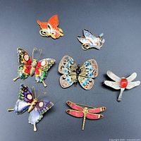 Top-down view of seven colorful butterfly and dragonfly brooches on black background showing various colors and metal tones.