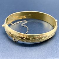 Side angle of gold-filled hinged bangle bracelet showcasing the engraved floral and leaf design on exterior surface and the attached safety chain.