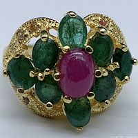 Close-up front view of the ring showing the oval cabochon ruby center stone surrounded by oval emeralds in a floral arrangement with small sapphires on the gold-plated silver band.