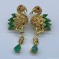 Front view of pair of bird motif earrings showing pear-cut emerald gemstone tails, dangling emerald drops, and round ruby stone eyes and body accents, all set in yellow gold plated 925 sterling silver.