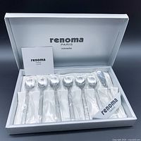 Set of six stainless steel spoons and one butter spreader arranged in a white Renoma Paris presentation box with logo visible inside lid.