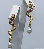 Pair of gold-plated sterling silver snake-shaped earrings with red round ruby stones and white dangling pearls displayed on a white stand