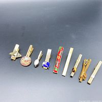 Nine vintage gold-tone tie clips displayed side-by-side on gray background, showing variety in design and size.