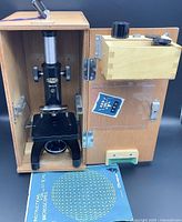 Full view of Olympus Tokyo microscope inside light-colored wooden carrying case. Microscope is upright with focusing knobs on sides, labeled Olympus Tokyo with serial number. A blue Olympus instruction manual lies in front of the case.
