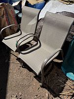 Two beige metal framed patio arm chairs with beige fabric seats and backs. Fabric has subtle square pattern. Chairs show normal outdoor age wear.