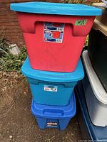 Stacked view of three plastic storage totes with lids in red, teal, and blue colors showing normal exterior wear.