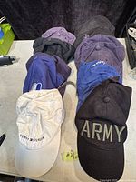 Photo showing ten baseball caps arranged in rows with the two branded caps visible at front - Tommy Hilfiger cream cap and black 'ARMY' cap. Other caps are mostly purple, blue, and black in color.