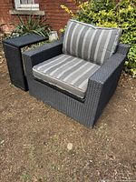Large black resin wicker outdoor armchair with gray and white striped cushions placed outdoors on dirt ground with plants and brick wall background.