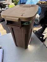 Front and side view of brown Rubbermaid Roughneck trash can with lid and handle, showing overall size and condition with some surface wear.