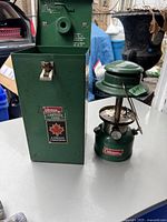 The green metal Coleman lantern next to its original green metal case with a latch and Canadian label.