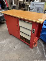 Full view of vintage metal workbench showing wood top, red frame, drawers, storage cabinet, and electrical outlet with orange cord.