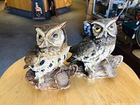 Two ceramic owl figurines sitting on a wooden table, showing their detailed painted features and textured bases.