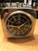 Front angle showing entire clock with polished silver-tone rounded square case, black dial with Roman numerals, and clock hands.