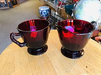Side view of the ruby red creamer and sugar bowl on a wooden surface showing handles and pedestal bases.