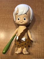 Photo of Bam Bam toy doll standing upright on wooden surface showing front view with green club and brown outfit
