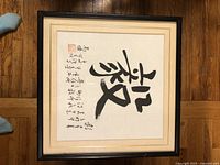 Image of first framed piece of Chinese calligraphy with red stamps and script, black modern frame, beige matting, on wooden floor.