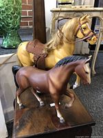 Two vintage horse toys on display, one tan with saddle and bridle, the other smaller and brown, both standing on a wooden surface