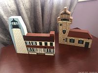 Two wooden lighthouse block models displayed on a table with curtain background. One model has a white lighthouse tower, brown roof building with shutters; the other model shows a stone lighthouse tower with attached building and brown roof.