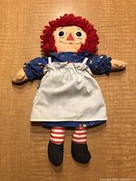 Front view of the 12 inch vintage Raggedy Ann doll showing the entire body with blue floral dress, white apron, striped legs, and black feet on a wooden surface.